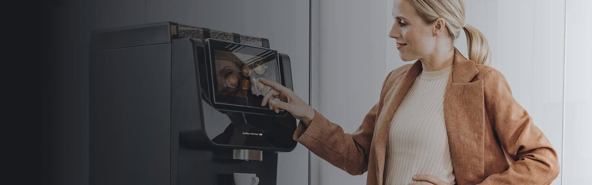 Eine Frau in eleganter Bürokleidung bedient einen Kaffeevollautomaten von Kaffee Partner im Büro. Eine weitere Frau ist im Vordergrund leicht unscharf zu sehen.