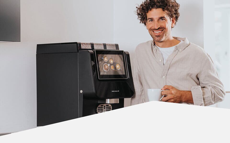 Mann steht mit Kaffeetasse lächelnd neben einem Kaffeevollautomaten