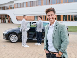 Eine lächelnde Vertrieblerin steht vor einem Auto und dem Kaffee Partner Gebäude, während im Hintergrund zwei männliche Kollegen im Gespräch sind. Sie trägt einen hellgrünen Blazer und strahlt Selbstbewusstsein aus.